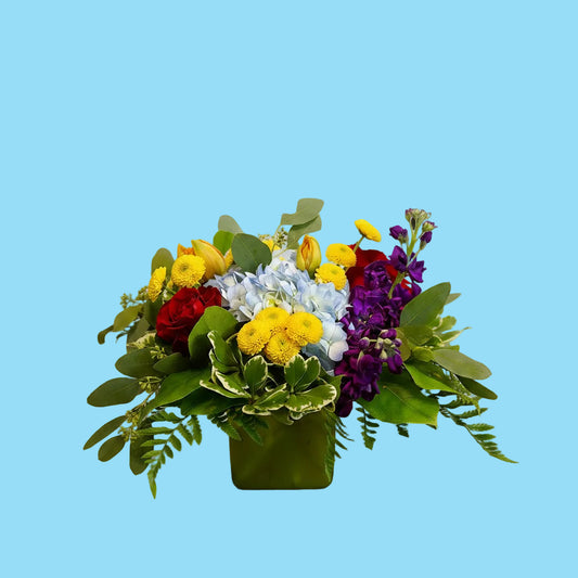 A vibrant mixed floral arrangement in a square green vase, featuring yellow daisy-like flowers, light blue hydrangeas, red roses, purple stock, and green foliage on a white background.